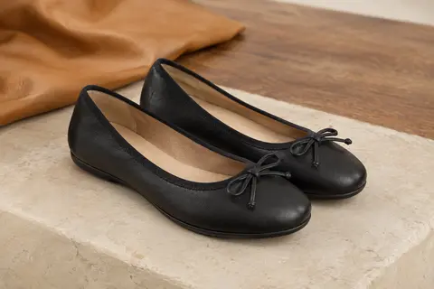Paire de ballerines noires à bout rond et petit nœud, présentées sur une pierre claire et du bois.
